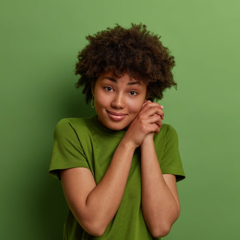 Pretty calm ethnic woman keeps hands together near face, looks optimistic at camera, dressed in casual t shirt, hears pleasant story from friend, isolated over green background. Monochrome shot